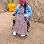 Plus size white woman sat in wheelchair wearing maxi floral dress, denim jacket, trainers and burgundy fedora hat
