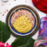a bowl of yellow thick pudding topped with nuts and pink petals on a gold tray staged with a multicoloured scarf.