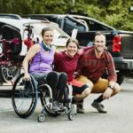 three people including one woman in wheelchair smiling side by side in a parking lot.