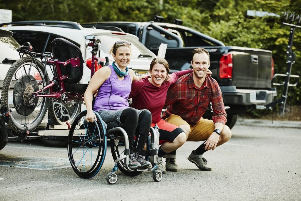three people including one woman in wheelchair smiling side by side in a parking lot.