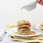 A stack of pancakes on a white plate with syrup being poured over top