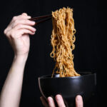 two hands holding a bowl of ramen noodles against a black background