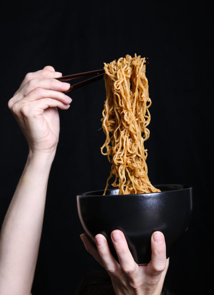 two hands holding a bowl of ramen noodles against a black background