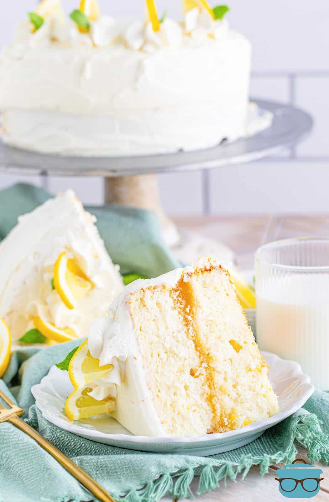 Slice of Lemon Layer Cake on white plate showing filling with whole cake in background on stand.