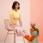 A model wearing a yellow short sleeved t-shirt and a pink checked skirt sits on a wire-framed chair and is admiring a pair of orange and yellow colourwork socks on her feet. Her expression is happy surprise