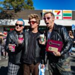 Three men wearing black jackets and sunglasses hold coffee cups and a plaque with a key on it in front of a 7-Eleven store.