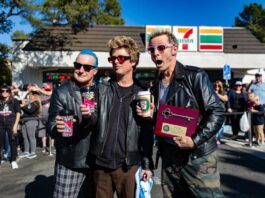 Three men wearing black jackets and sunglasses hold coffee cups and a plaque with a key on it in front of a 7-Eleven store.