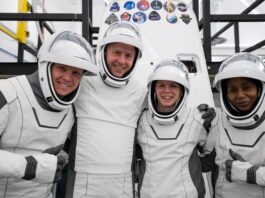 four people in white spacesuits with the visors up pose and smile in front of a white cone-shaped spacecraft